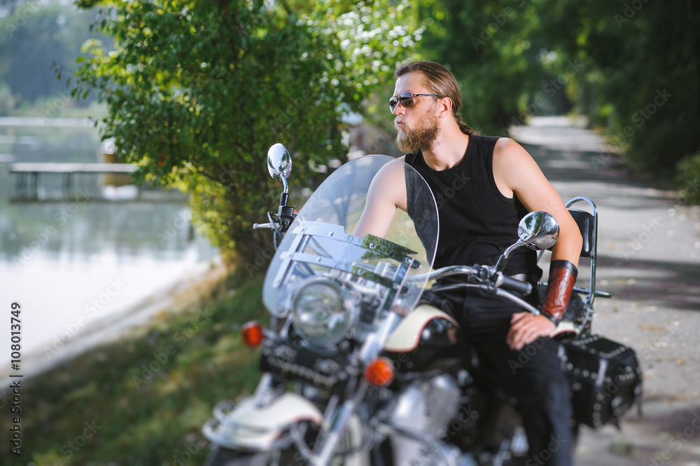 Handsome biker in sunglasses with long hair and beard sitting on his motorcycle near the lake on a sunny day. Tilt shift lens blur effect.