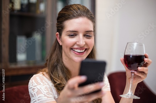 Cheerful woman holding wine glass while using phone