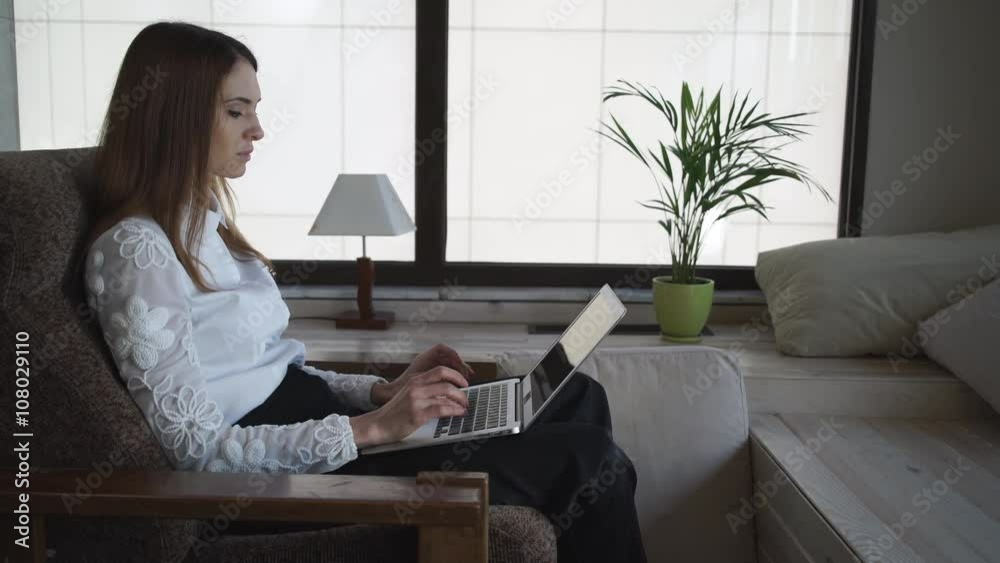 Woman with long hair at home sitting and working near panoramic windows . Her hands with short pink nails lying on the armchair. She is typing on the laptop. Female dressed in white shirt and black Woman with long hair at home sitting and working near panoramic windows . Her hands with short pink nails lying on the armchair. She is typing on the laptop. Female dressed in white shirt and black
