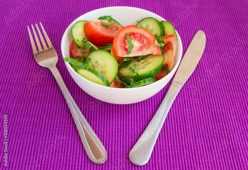 Fresh spring vegetable salad (cucumbers, tomatoes) in a white bowl on a bright purple background