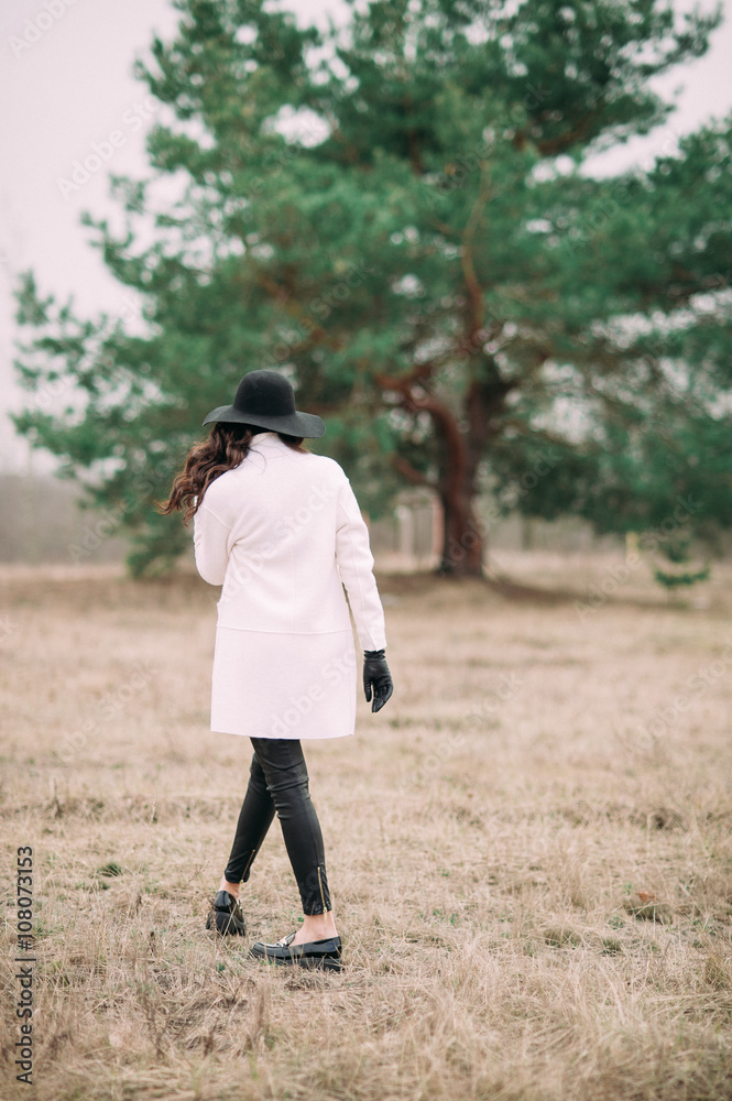 Beautiful fashionable woman in a black hat and white coat posing outdoors