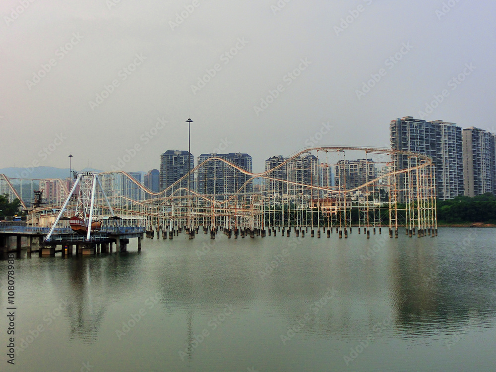 Abandoned Chinese amusement park across water with old rollercoaster and pirate ship ride - landscape color photo