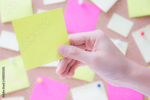 Hand holding empty post it paper or sticky note with blurred cork board background for insert your messages.
