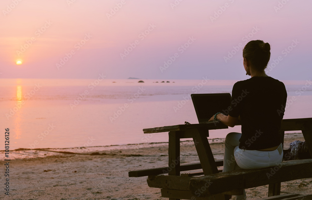 Young woman sitting at the table with a laptop in front of sunset view. Lady frelanser working at the beach.

