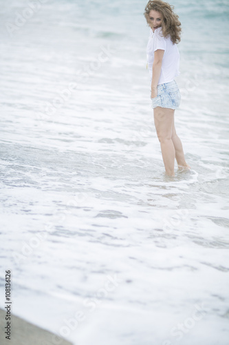 enter into the water. Girl with wavy hair on the beach with black sand in a light white top and denim varieties