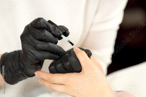 Manicurist cover colorless varnish nails client. Manicure master in black gloves does manicure