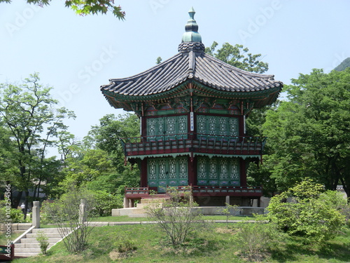 A two storey wood built Pagoda style building in Seoul, South Korea. Grass and bushes in the foreground, trees in the background with a clear blue sky.