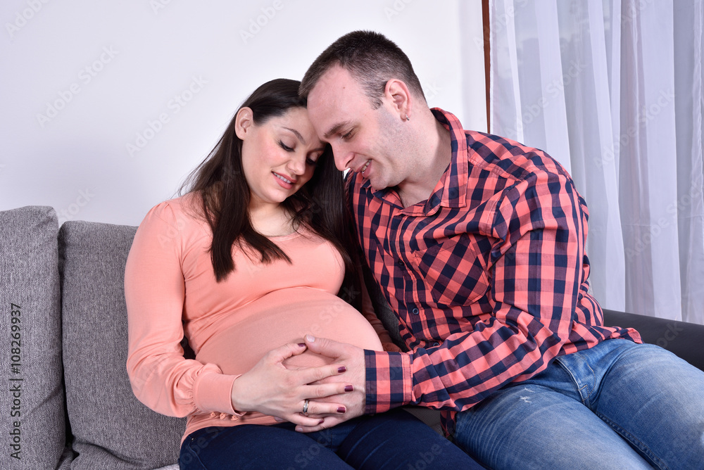 Young smiling couple in love expecting a baby. They are sitting on the sofa and enjoying.