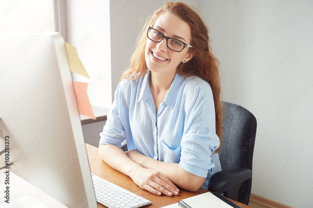 Attractive young female entrepreneur with red hair and spectacles looking and smiling at the camera during small break, in front of the computer while working in the office. Business concept Attractive young female entrepreneur with red hair and spectacles looking and smiling at the camera during small break, in front of the computer while working in the office. Business concept