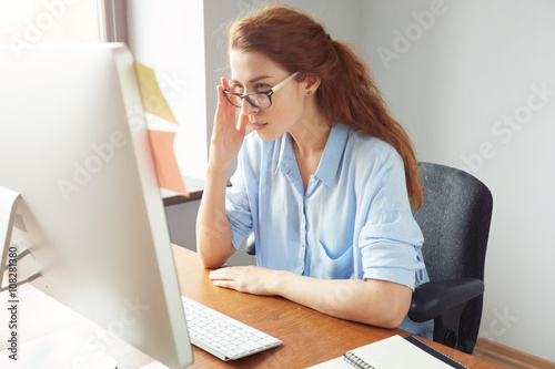 Attractive thoughtful redhead businesswoman looking at the screen with concentrated expression in the office. Portrait of female freelancer in glasses and blue shirt working on the laptop © wayhome.studio 