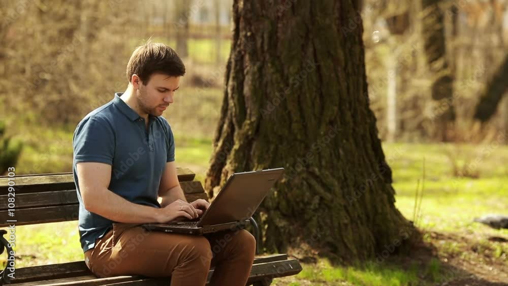 Young Man Sitting On Park Bench Using Laptop Young Man Sitting On Park Bench Using Laptop
