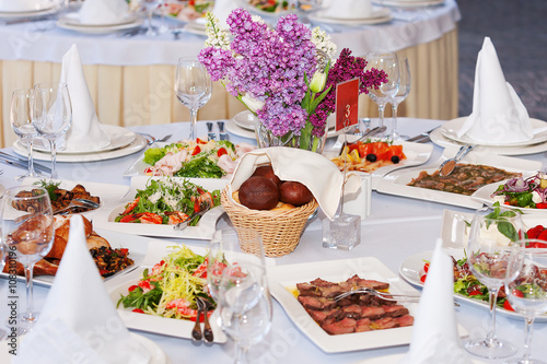 Table set for banquet with salads, bread, roasted meat. Flower decoration.