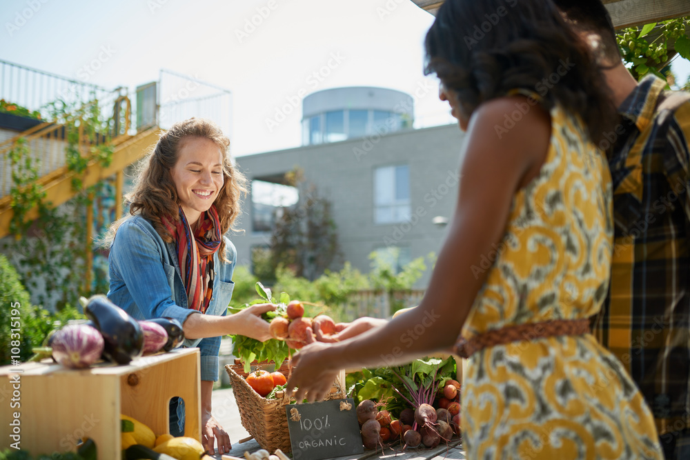 Friendly woman tending an organic vegetable stall at a farmer's market and selling fresh vegetables from the rooftop garden - obrazy, fototapety, plakaty 