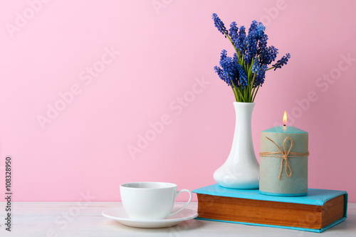 Blue muscari flower in a beautiful white vase on a wooden table with a candle on a book and a Cup on a saucer on a pink background