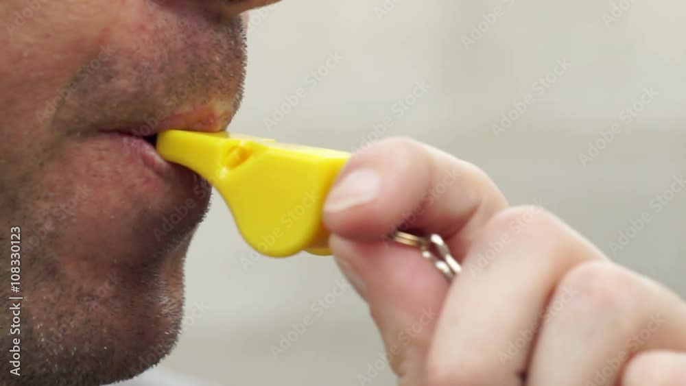 Extreme close up shot of a man blowing on a yellow whistle to make noise, give warning, get someone's attention or signal that something important is happening.