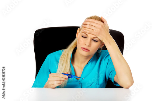 Tired young female doctor or nurse sitting behind the desk and holding termomether photo