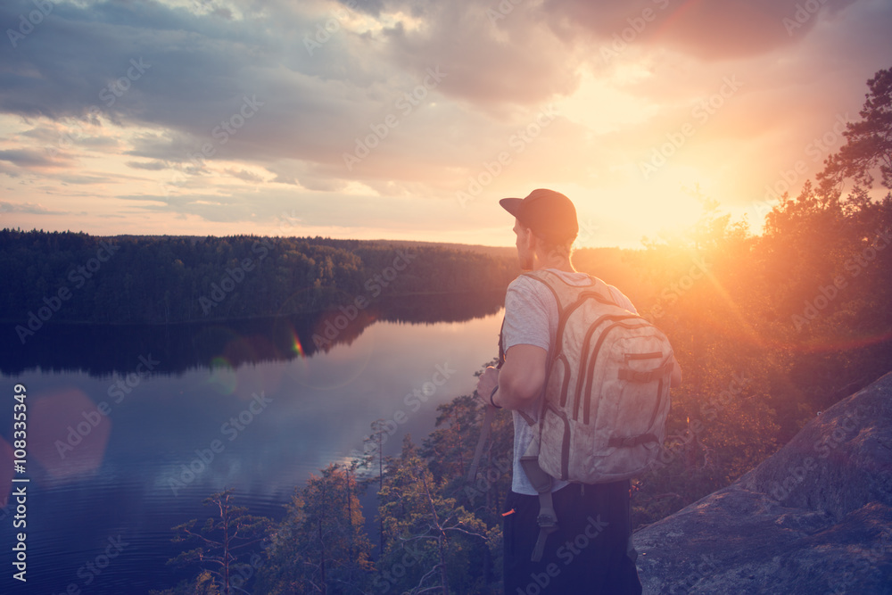 Young casual man with backpack and cap on the cliff above river and looking far away (intentional sun glare and lens flare)