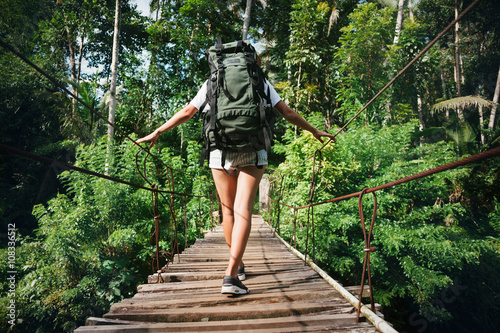 Quadro em tela Young woman with backpack traveling across hanging bridge in tropical forest
