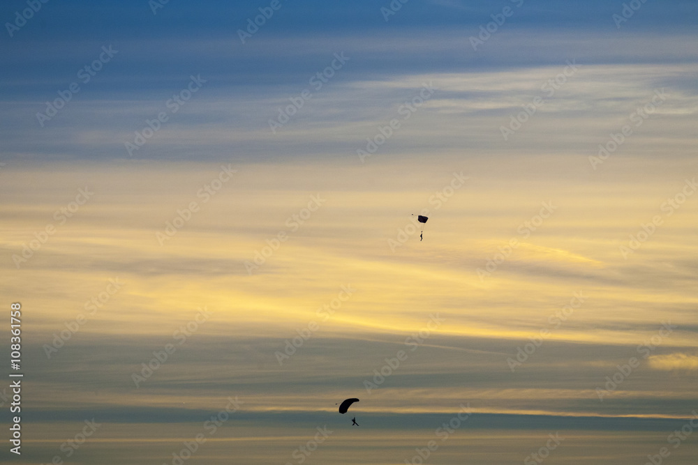 Silhouette of two skydivers falling with parachute open against dramatice sunset and clouds