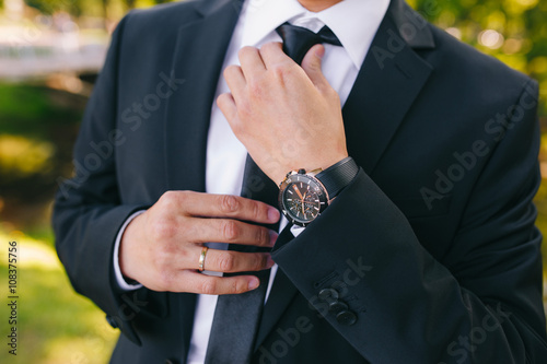 A man in a black suit straightens his tie. A stylish watch on your hand