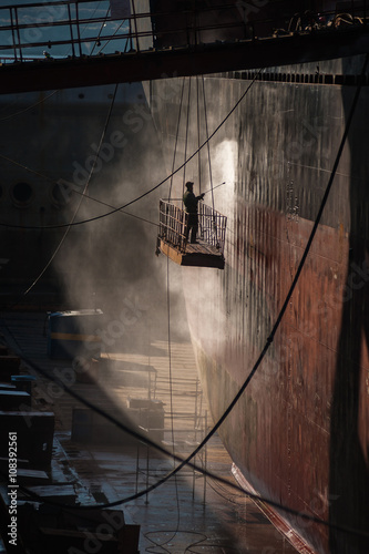 Shipyard worker power washing a ship on dry dock.