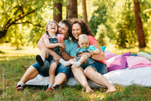 Young happy family with kids having picnic with colored pillows outdoors. Parents with two children relax in a sunny summer garden. Mother, father, little girl and baby boy playing in park.