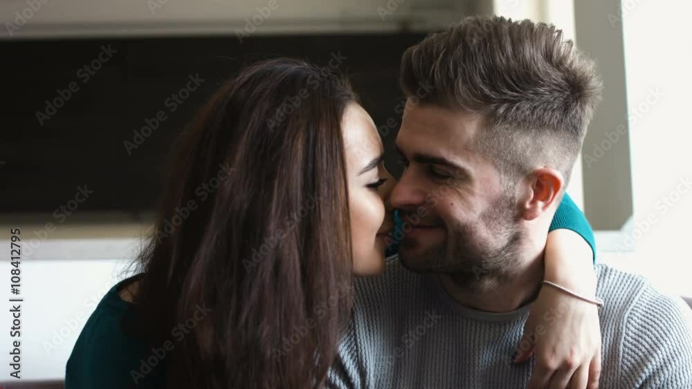 Beautiful couple sitting on a sofa, young man checking his emails in laptop and talking to girlfriend , kissing her on the lips. Beautiful couple sitting on a sofa, young man checking his emails in laptop and talking to girlfriend , kissing her on the lips.