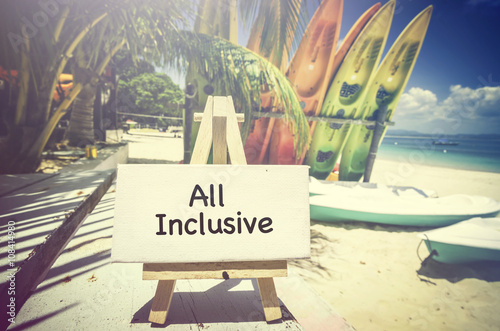 conceptual image with word ALL INCLUSIVE on white canvas frame and wooden tripod. Blurred image of beach and colorful kayaks background.