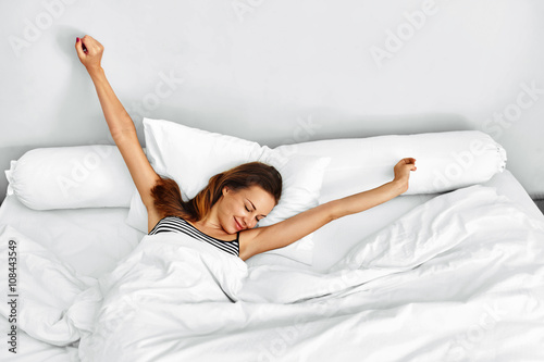 Morning Wake Up. Smiling Young Woman Waking Up Fully Rested On White Bedding. Model Stretching In Bed. Girl Lying, Relaxing In Bedroom. Healthy Sleep, Lifestyle. Wellness, Health, Beauty Concept