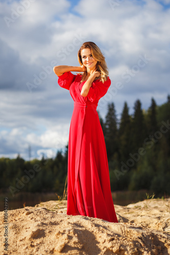 Gorgeous red head young woman in long red dress on a background of a lake. Sandy canyon. Fashion style