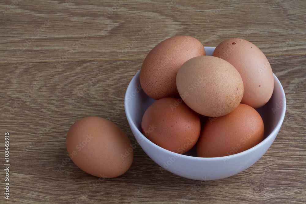 A white bowl full of fresh, brown, speckled eggs on a wooden table. A white bowl full of fresh, brown, speckled eggs on a wooden table.