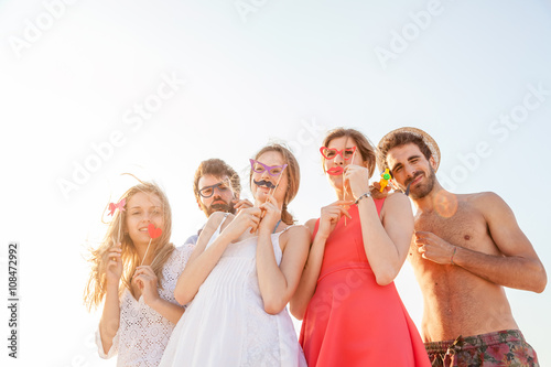 Summer, sandy beach at sunset. A group of friends at a party on the beach, having fun with mustaches, hearts, fake glasses and objects to joke