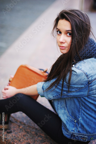 beautiful girl in blue jeans jacket and scarf sitting on a granite parapet. In her hand brown elegant handbag