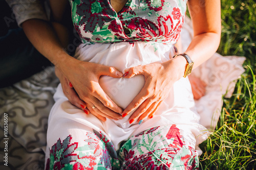 Pregnant Woman and Her Husband holding her hands in a heart shape on her baby bump.