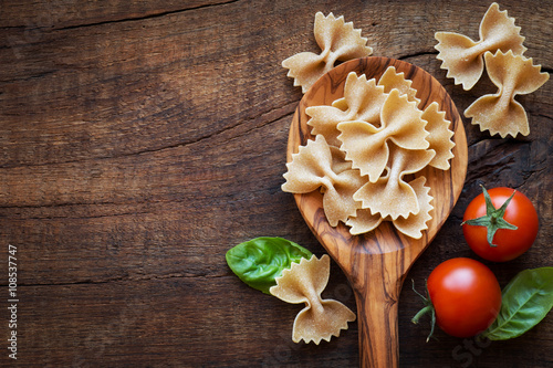 Italian cuisine concept - Wholemeal farfalle pasta in wooden spoon with fresh basil and tomatoes od dark rustic wooden background . With copy space for your text
