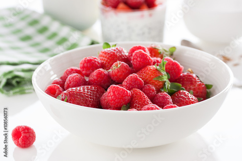 Summer fresh red juicy fruits – raspberries and strawberries in a white bowl on a table for breakfast or lunch, closeup, selective focus.
