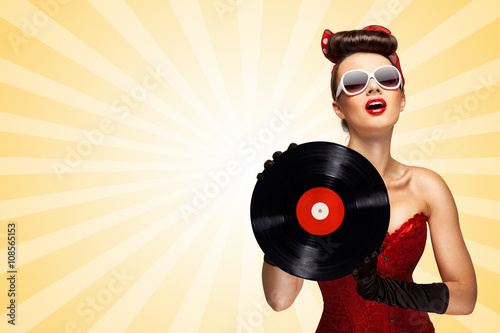 Retro play / Vintage photo of glamorous pinup girl wearing long gloves and dressed in a red sexy corset, holding LP vinyl record on colorful abstract cartoon style background.