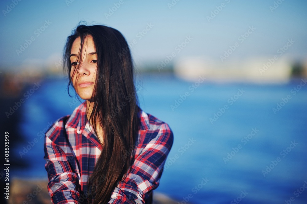 beautiful brunette girl in plaid shirt sitting on the embankment near the river