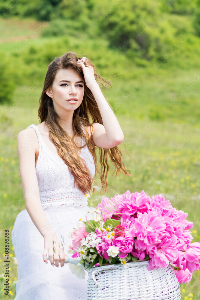 Gorgeous young boho fashion woman in white dress on bike with white basket with pink peonies Gorgeous young boho fashion woman in white dress on bike with white basket with pink peonies