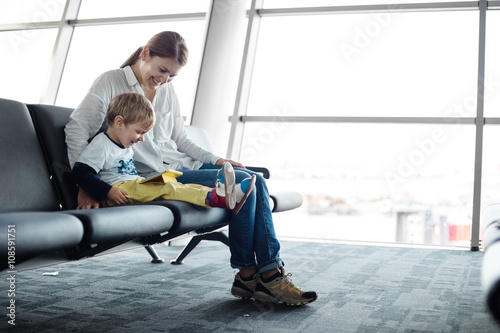 Little boy and his mother sitting in an airport departure hall and playing on a tablet 