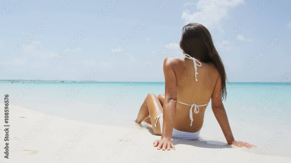 Beach vacation woman relaxing on sand happy in enjoyment under the sun. Asian mixed race model looking happy sitting on perfect white sand beach and turquoise ocean water on luxury summer vacation. Beach vacation woman relaxing on sand happy in enjoyment under the sun. Asian mixed race model looking happy sitting on perfect white sand beach and turquoise ocean water on luxury summer vacation.