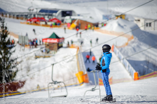 Side view of female skier standing on a ski slope at a sunny day with ski-lift and lively ski resort on the background. Winter sports concept.