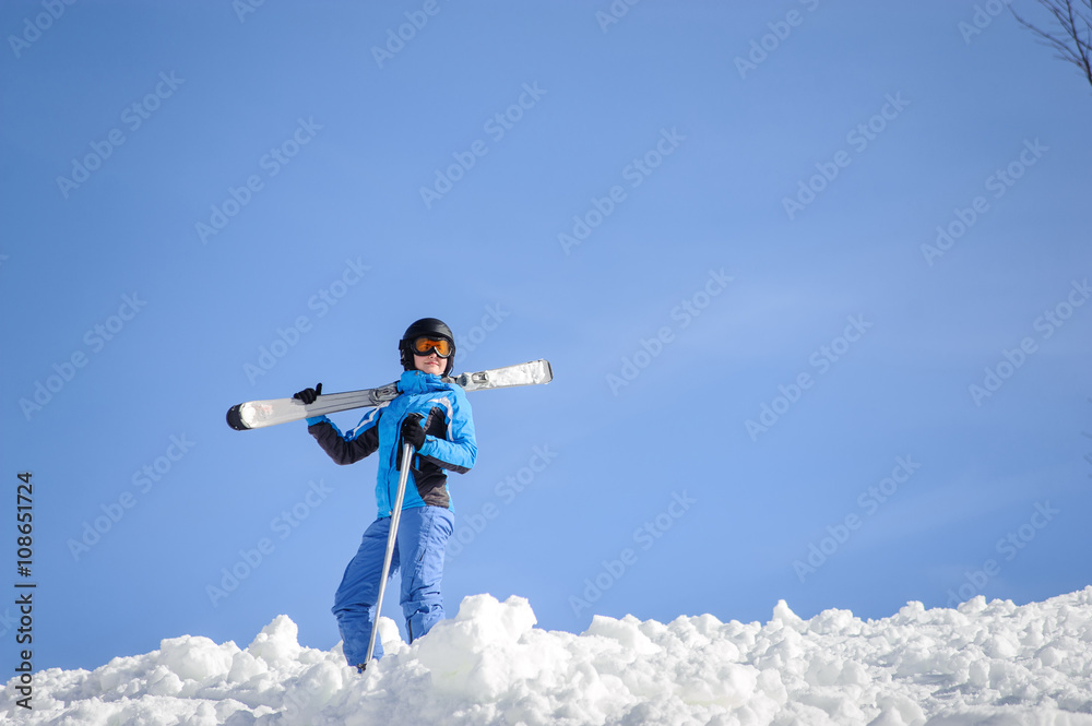 Portrait of young female skier standing on top of the mountain against blue sky on a sunny day. Woman is holding skis on her shoulder smiling and looking into the distance. Winter sports concept. Portrait of young female skier standing on top of the mountain against blue sky on a sunny day. Woman is holding skis on her shoulder smiling and looking into the distance. Winter sports concept.