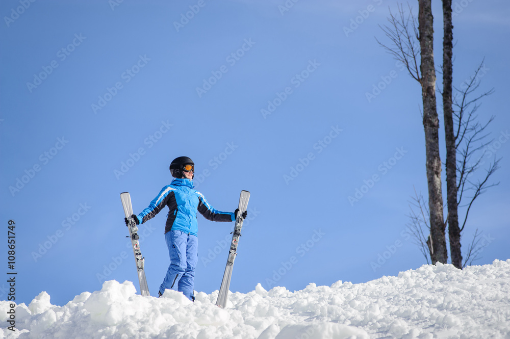 Young female skier wearing blue ski suit helmet and goggles on sunny day. Woman is holding her skis. Winter sports concept.