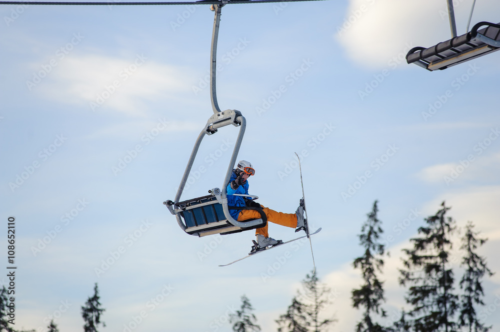 Skier sitting at ski lift against blue sky. Skier is wearing blue jacket, orange pants, helmet and orange goggles. Winter sports concept. Ski resort at Carpathian Mountains, Bukovel, Ukraine Skier sitting at ski lift against blue sky. Skier is wearing blue jacket, orange pants, helmet and orange goggles. Winter sports concept. Ski resort at Carpathian Mountains, Bukovel, Ukraine