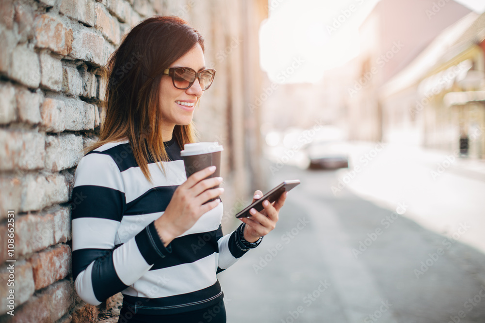 Young woman standing at the street drinking coffee to go and using mobile phone
