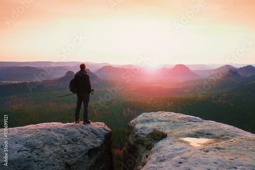 Hiker with sporty backpack stand on rocky view point above misty valley. Sunny spring daybreak