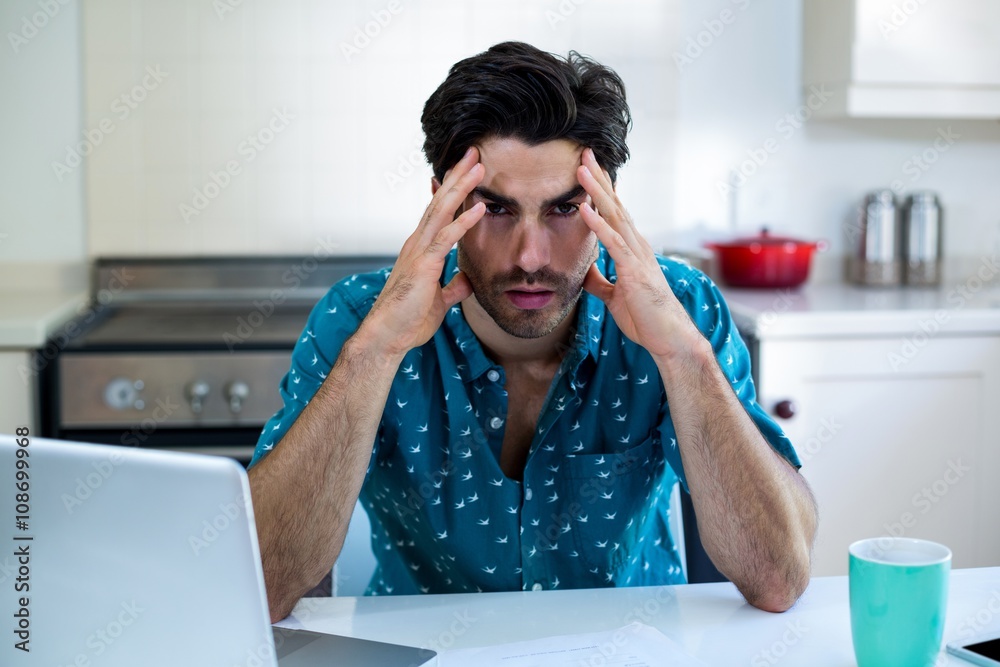 Tensed man sitting with bills and laptop in kitchen