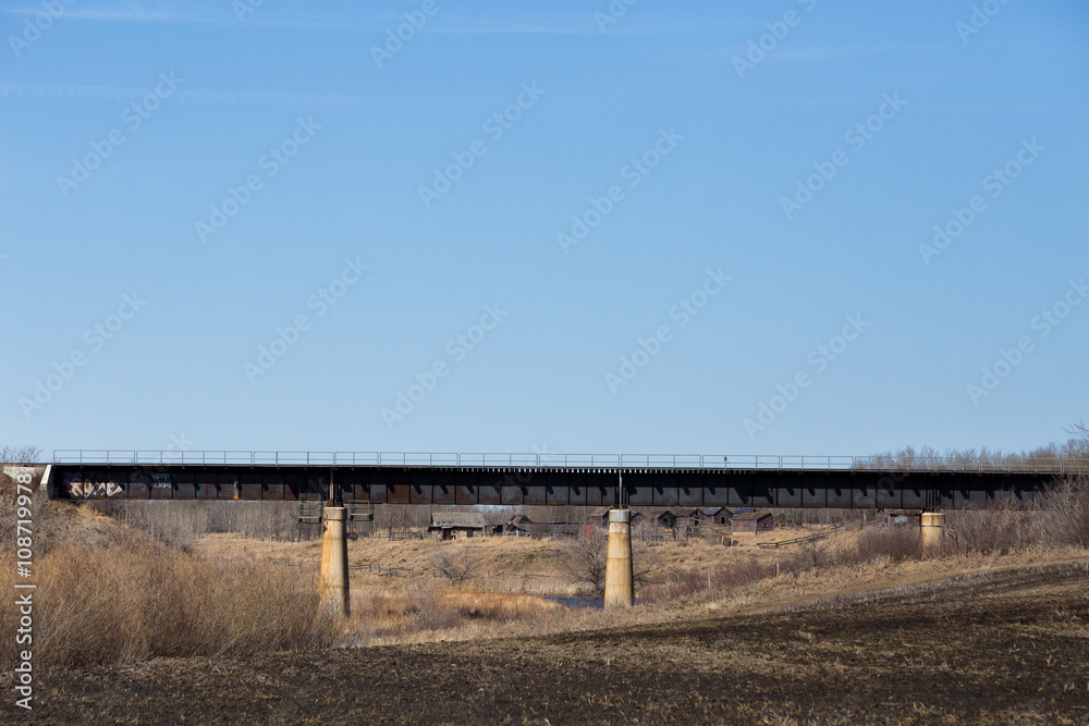 A steel railway bridge with concrete pillars over small valley with crumbling fence and buildings in spring countryside landscape