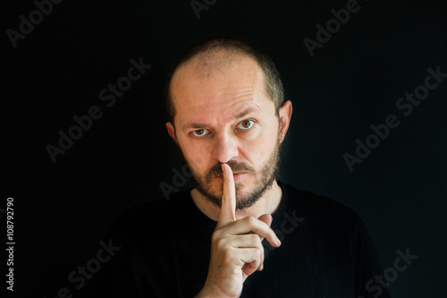 Serious man with beard and mustaches on black background in low key, making silence gesture, pst, shh